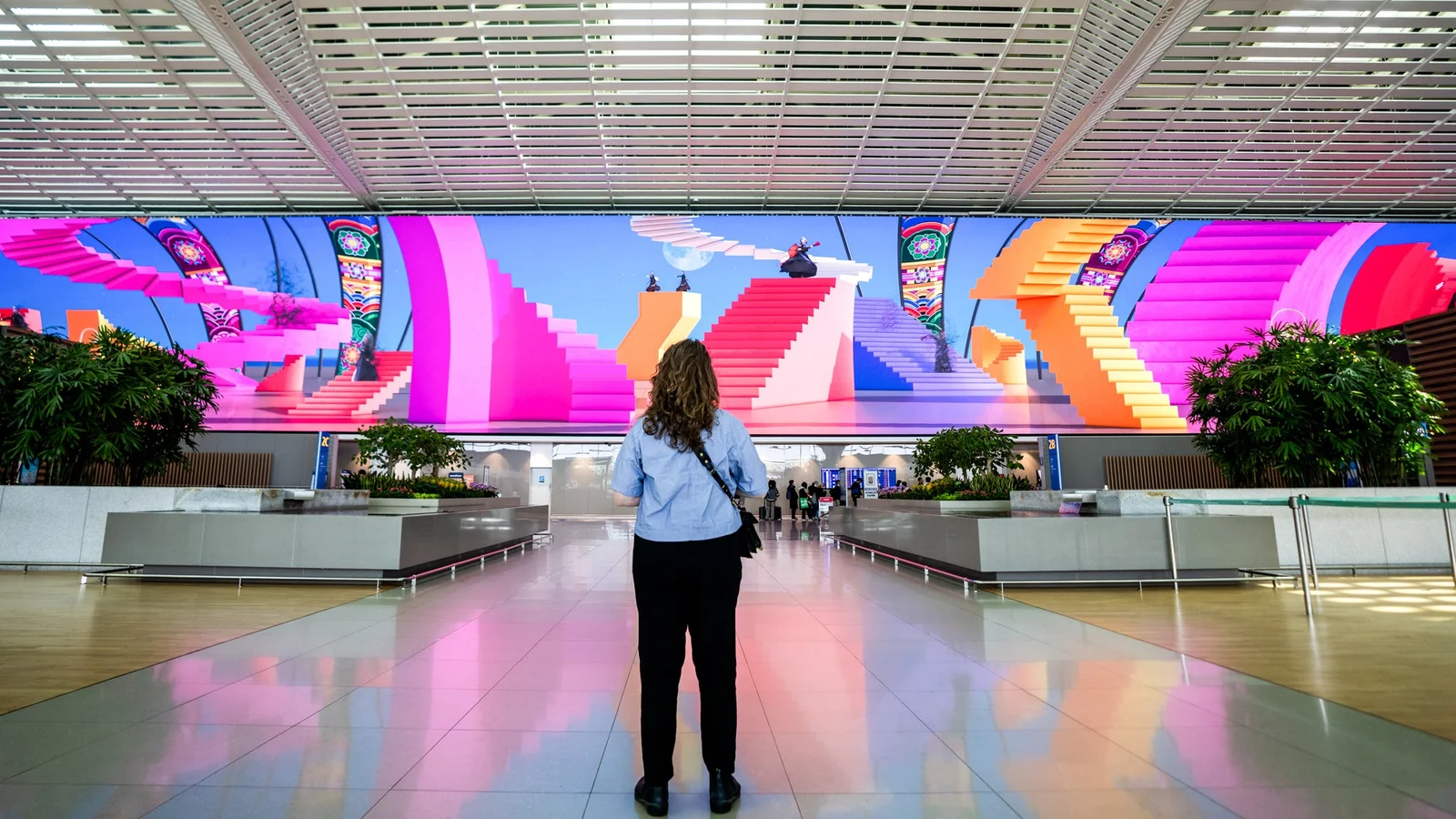 ソウル・仁川空港ターミナル2の16K大型LEDキャンバス(Moment Factory制作)──韓国の文化コンテンツカプセルの一場面、幾何学的なピンクの階段を描いた巨大な曲面ディスプレイの前に1人の旅客が立つ
